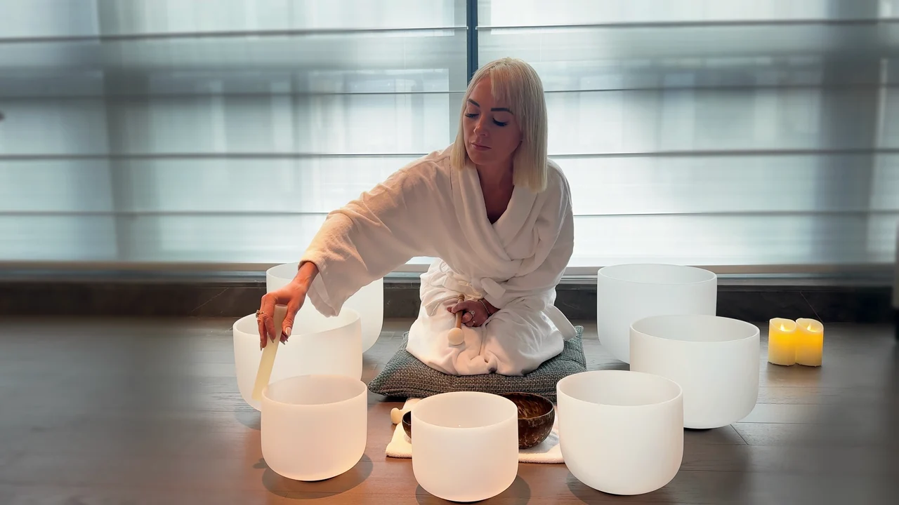 Viktoria in a white robe playing white crystal singing bowls beside candlelight.