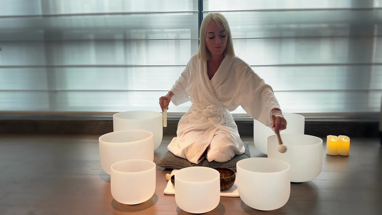 Viktoria in a white robe playing white crystal singing bowls beside candlelight.